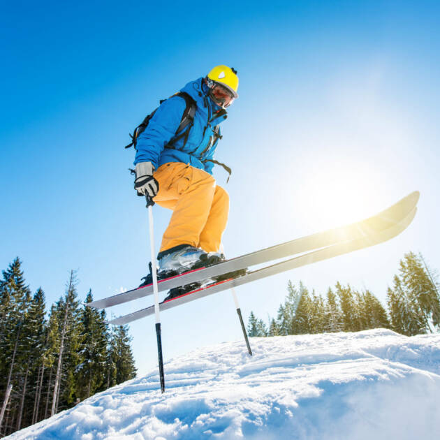 Low angle shot of a skier jumping in the air while skiing in the mountains copyspace. Blue sky, sun and winter forest on the background